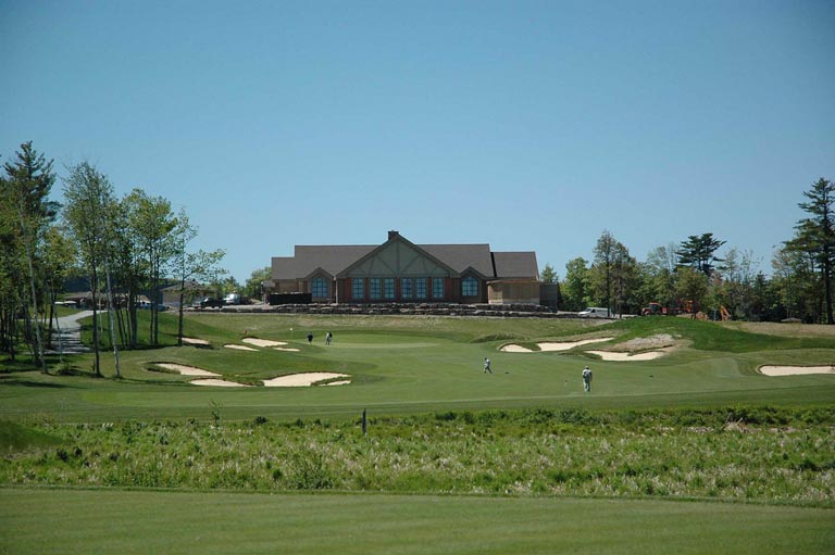 Colour photograph of green grass with spots white sand and a building behind