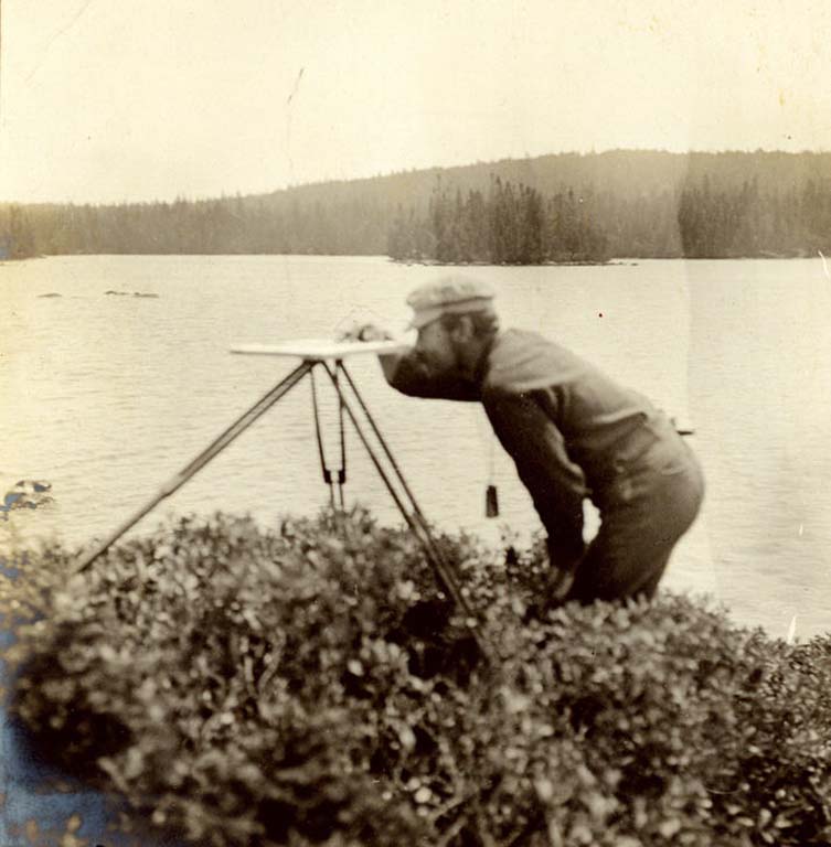 Photographie noir et blanc d’un homme devant un lac avec de l’équipement d’arpentage