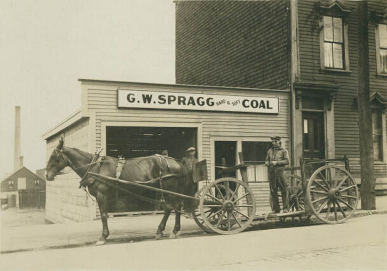 Black and white photograph of a horse and wagon with three men in front of a wooden building